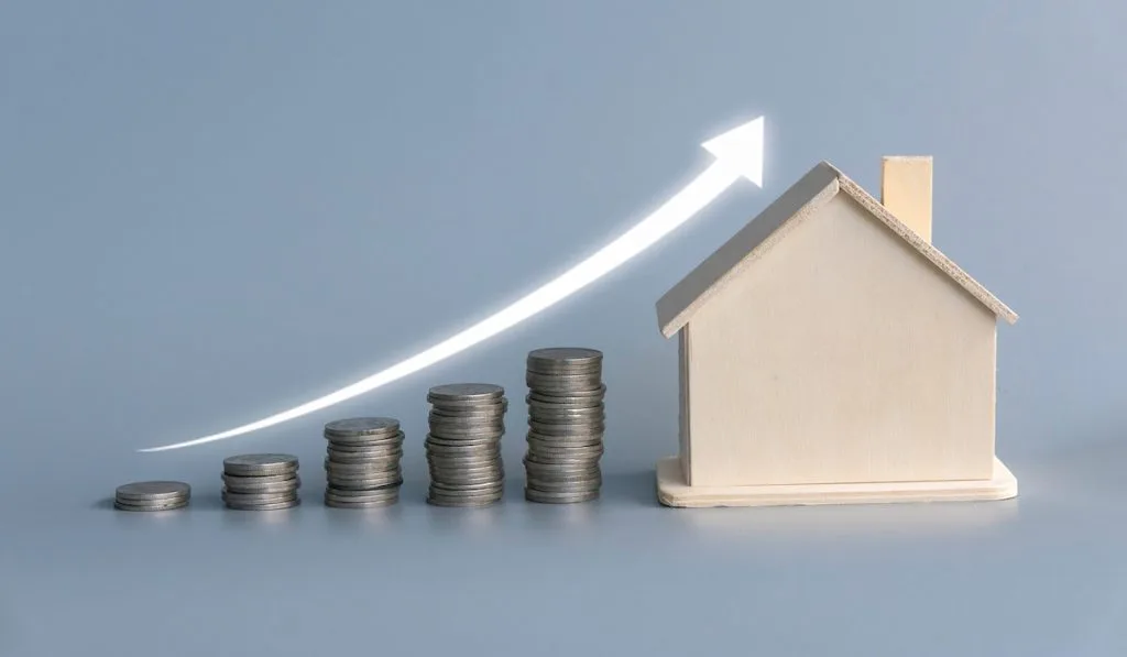 A stack of ascending coins with house model and glowing white arrow on grey background.
