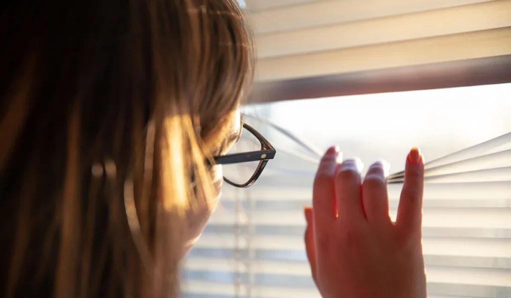 A woman looks through the blinds at the early morning sunlight.
