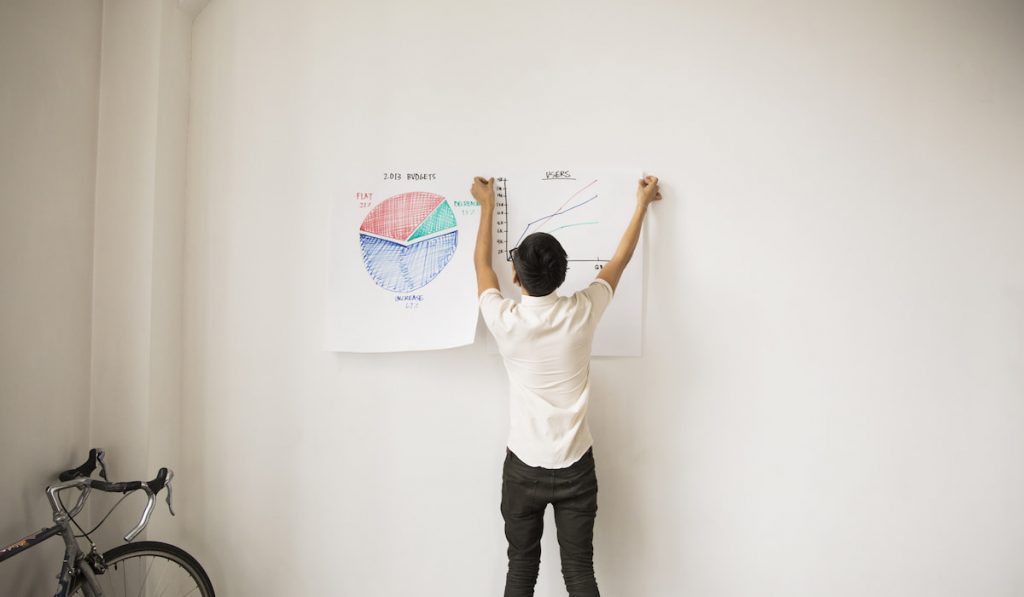 Rear View Of Businessman Sticking Charts On White Wall 