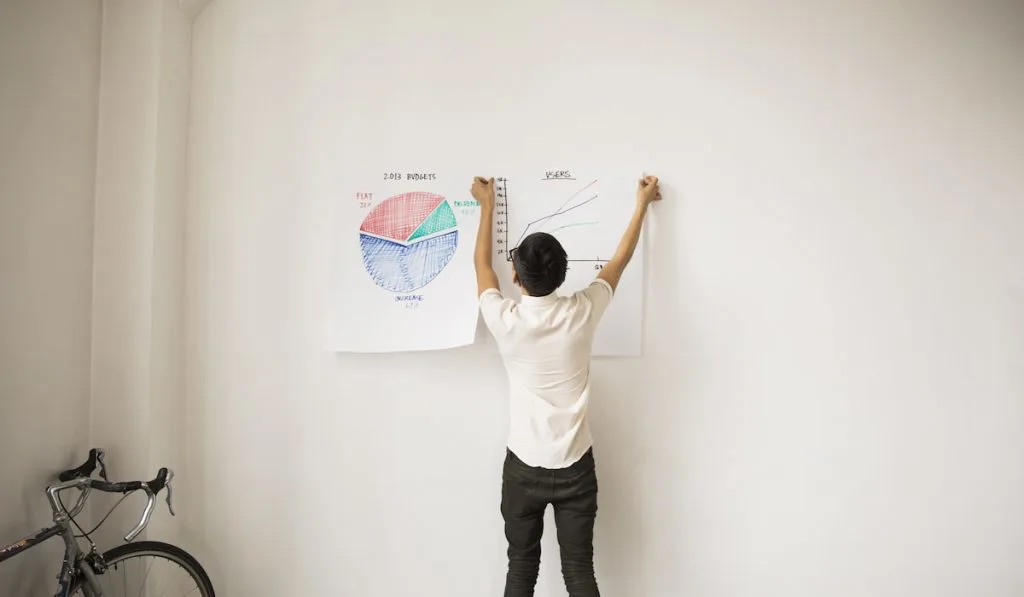 Rear View Of Businessman Sticking Charts On White Wall 